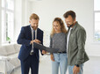 Happy married couple buying new house. Young husband and wife standing in white living room of partially furnished apartment and looking at contract that their real estate agent gives them to sign